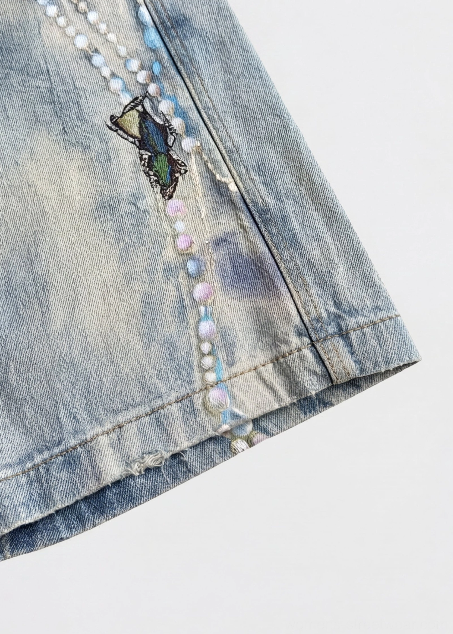 Side hem close-up of acid wash blue denim jorts with pearl chain butterfly embroidery and raw frayed edge