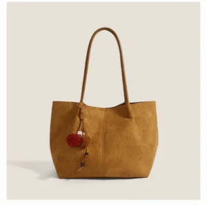 Caramel brown faux suede pom charm tote bag with dual shoulder straps and decorative yarn pom pom tassel on a minimalist studio background