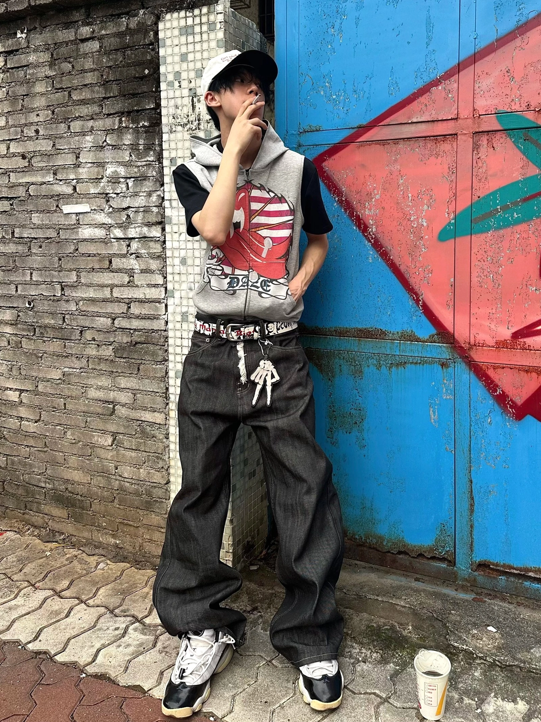 Male model wearing Americana lips graphic sleeveless zip hoodie vest with baggy black jeans at graffiti brick wall