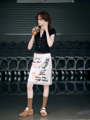 Apricot cream kitty graffiti midi pencil skirt styled with a black polo shirt, brown socks, and sneakers in a casual streetwear setting
