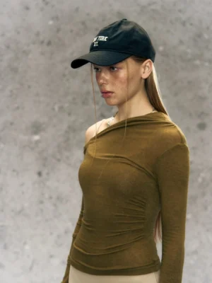 Olive army green asymmetrical one-shoulder long sleeve streetwear top styled with a black baseball cap
