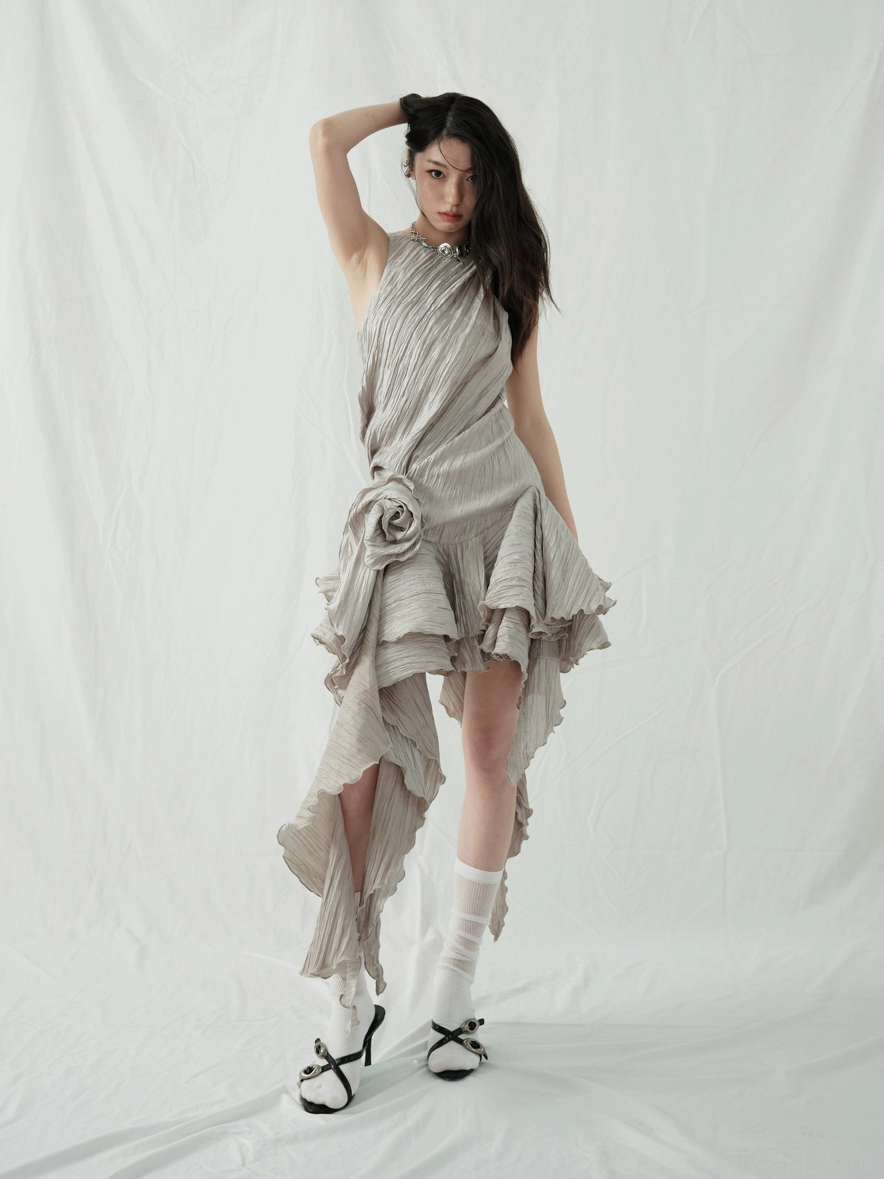 Back three-quarter view of an ash asymmetric metallic party dress with layered high-low ruffle hem, crinkled fabric, white crew socks, and bow detail heels