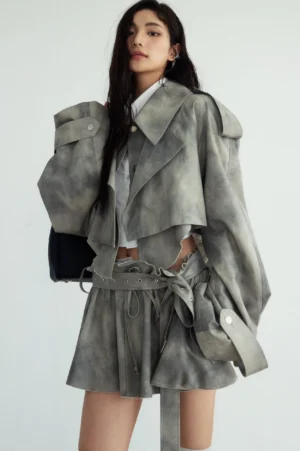 Front three-quarter view of an ash gray cropped trench jacket and ruffled mini skirt streetwear set with notched lapels, shoulder epaulets, faded tie-dye texture, and belted waist detail