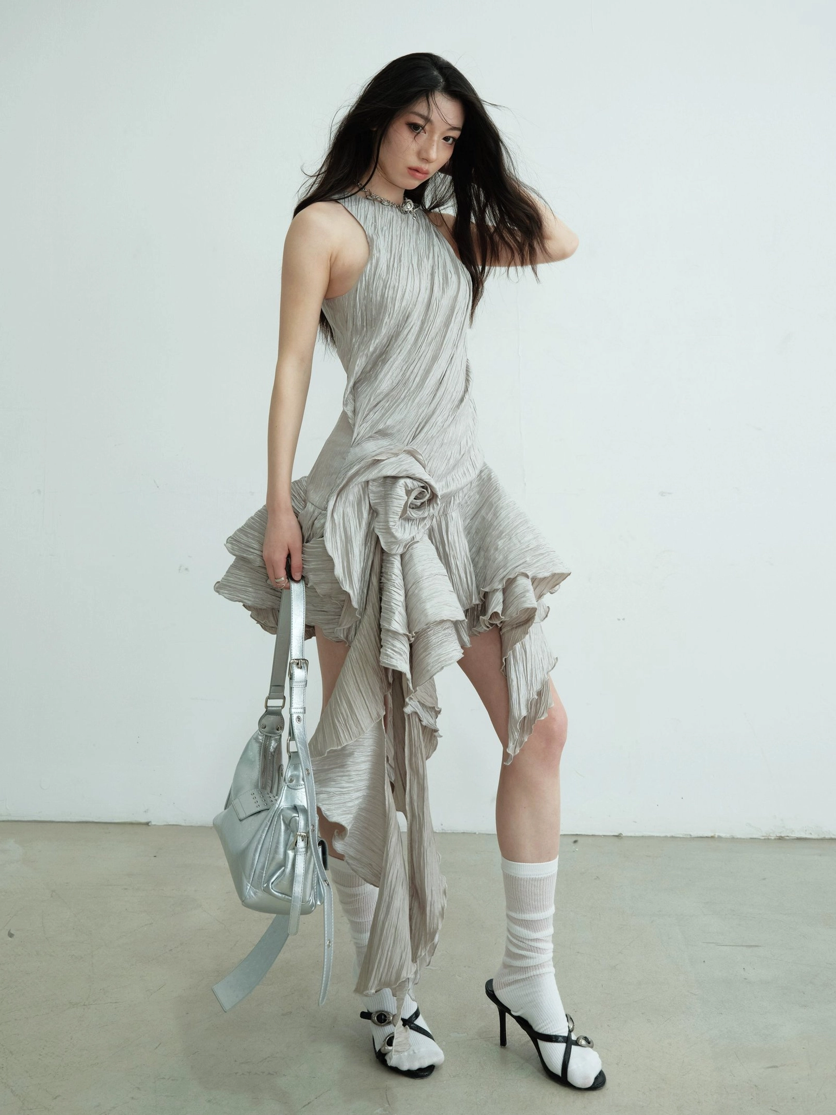 Front view of an ash halter neck metallic party mini dress with crinkled texture, asymmetric ruffle hem, silver shoulder bag, and bow heels