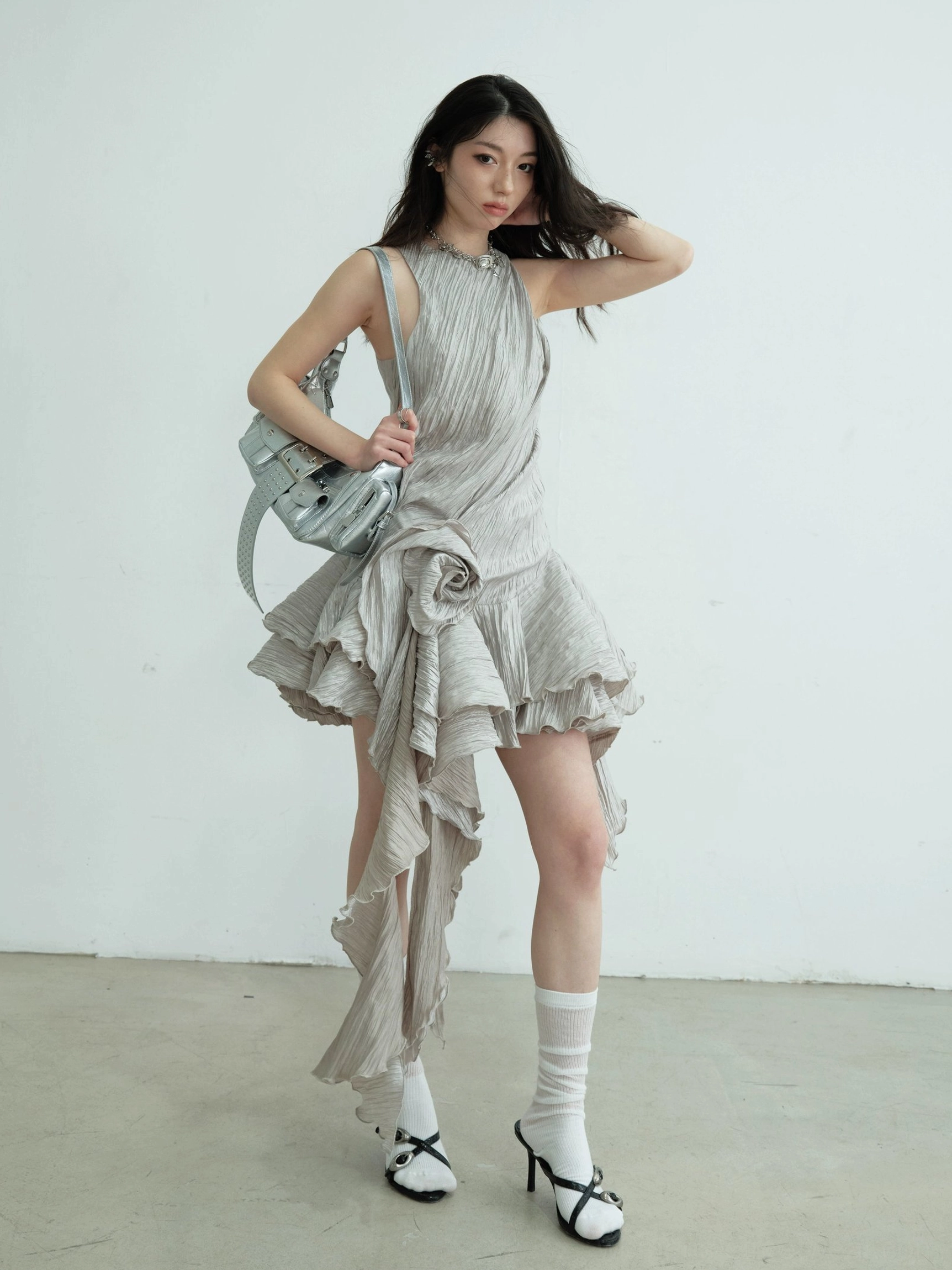Full front view of an ash metallic party mini dress with sleeveless halter cut, crinkled woven texture, asymmetric ruffle hem, floral ornament, and silver shoulder bag