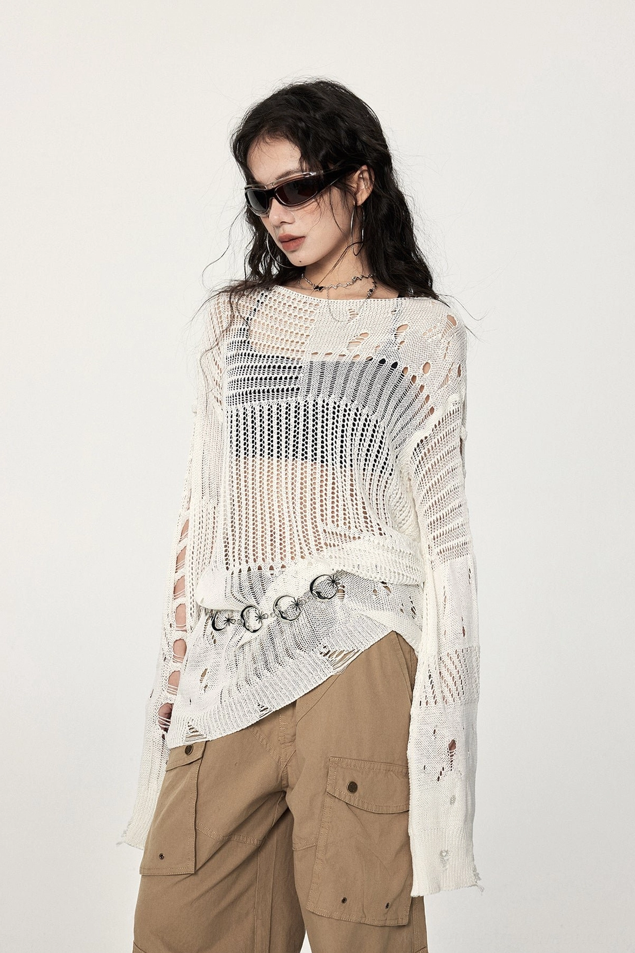 Low-angle view of an asymmetric distressed crochet sweater with slit long sleeves, open knit fabric, metal waist rings, and urban streetwear styling