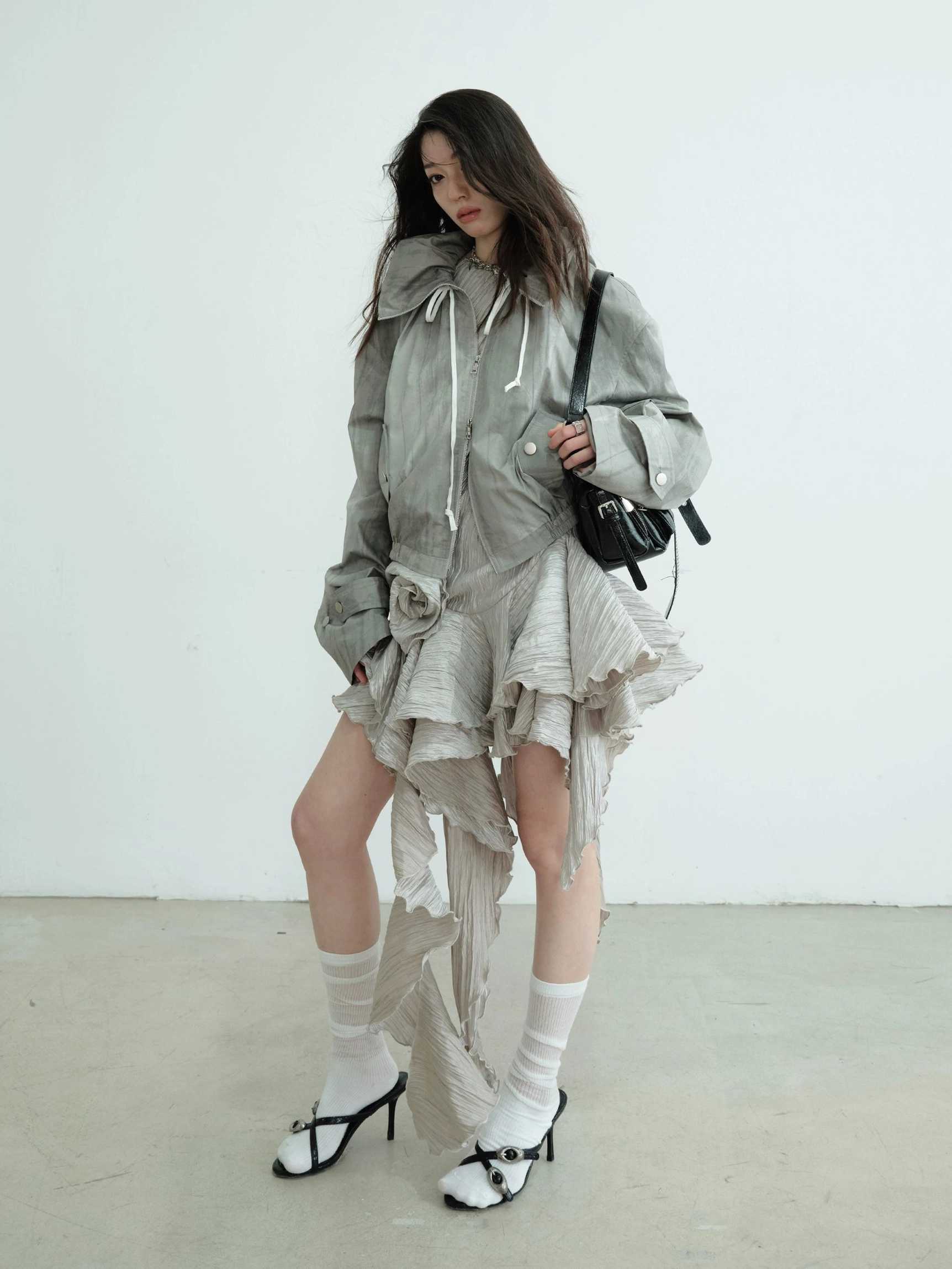 Side styling view of an asymmetric metallic ruffle hem party mini dress layered with an oversized gray hooded jacket, black crossbody bag, white socks, and bow heels