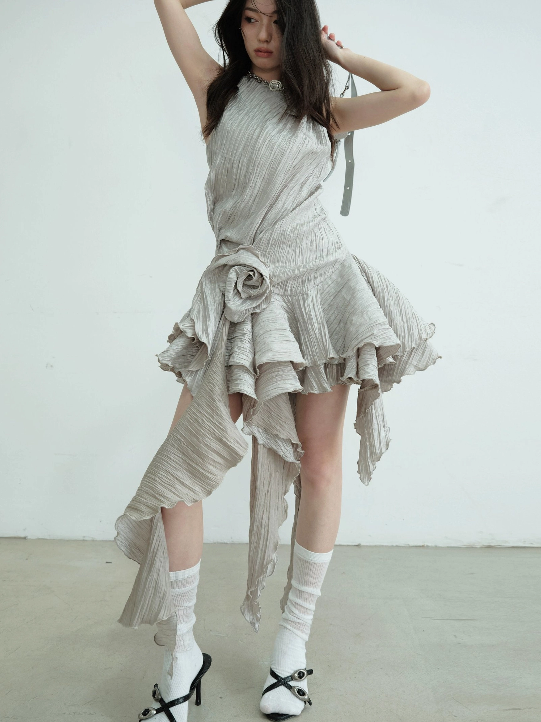 Low-angle front side view of an asymmetric metallic ruffle hem mini dress with three-dimensional floral decoration, crinkled texture, and silver shoulder bag