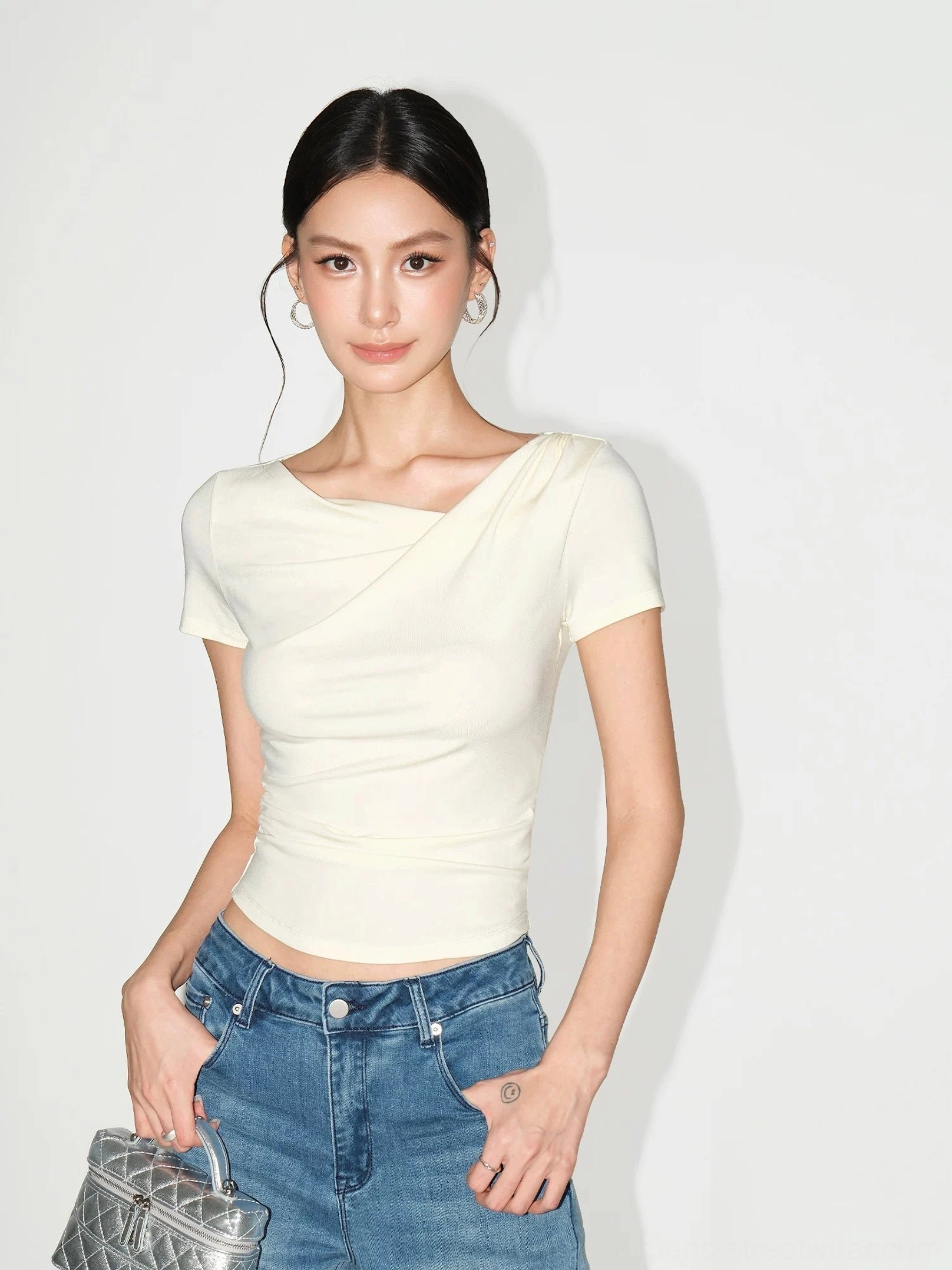 Cream asymmetric off-shoulder ruched slim fit top styled with blue denim jeans in a minimalist studio