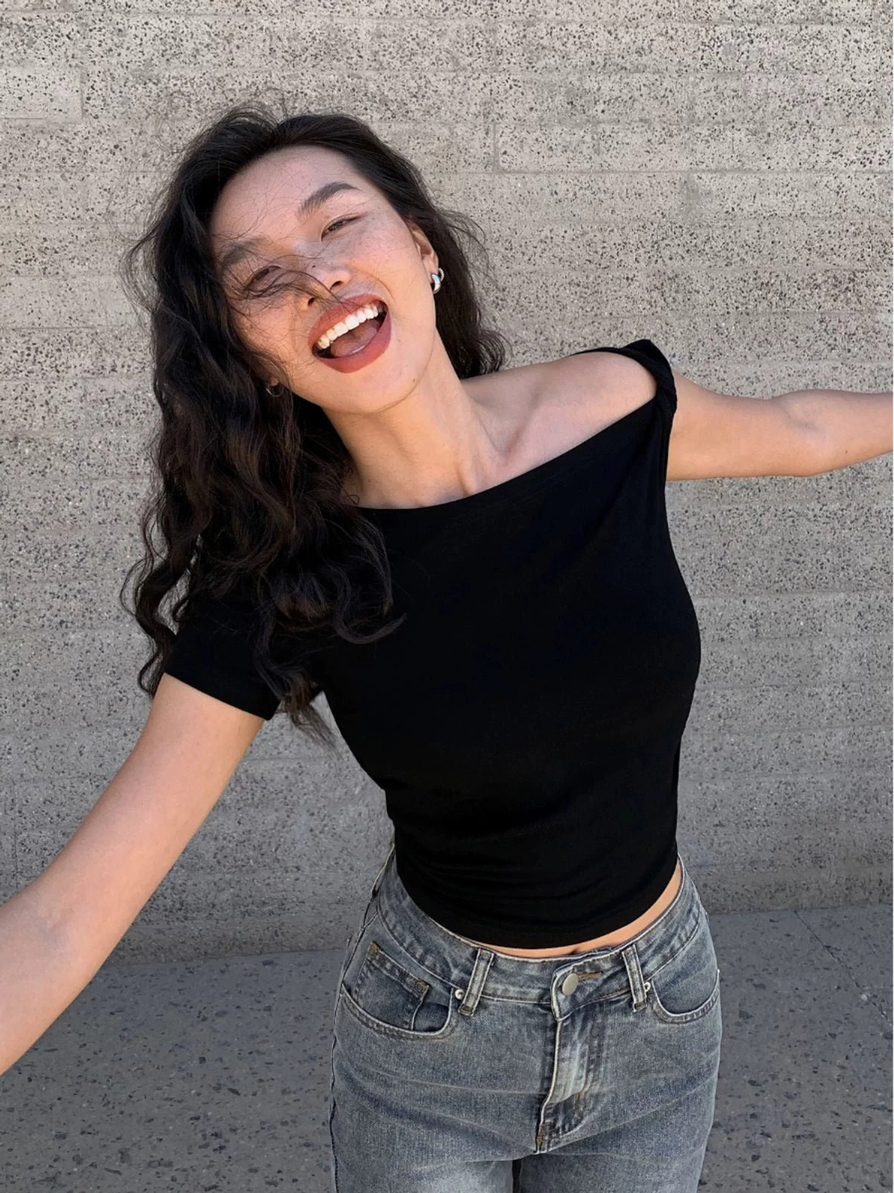 Black asymmetric one-shoulder cropped tee with a slim fitted silhouette styled with blue jeans against an outdoor concrete wall