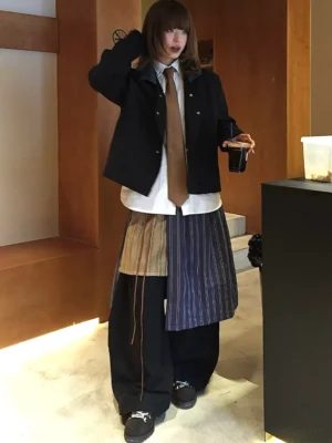 Front full-body view of asymmetric pinstripe apron balloon streetwear pants with black wide legs and layered contrast panels