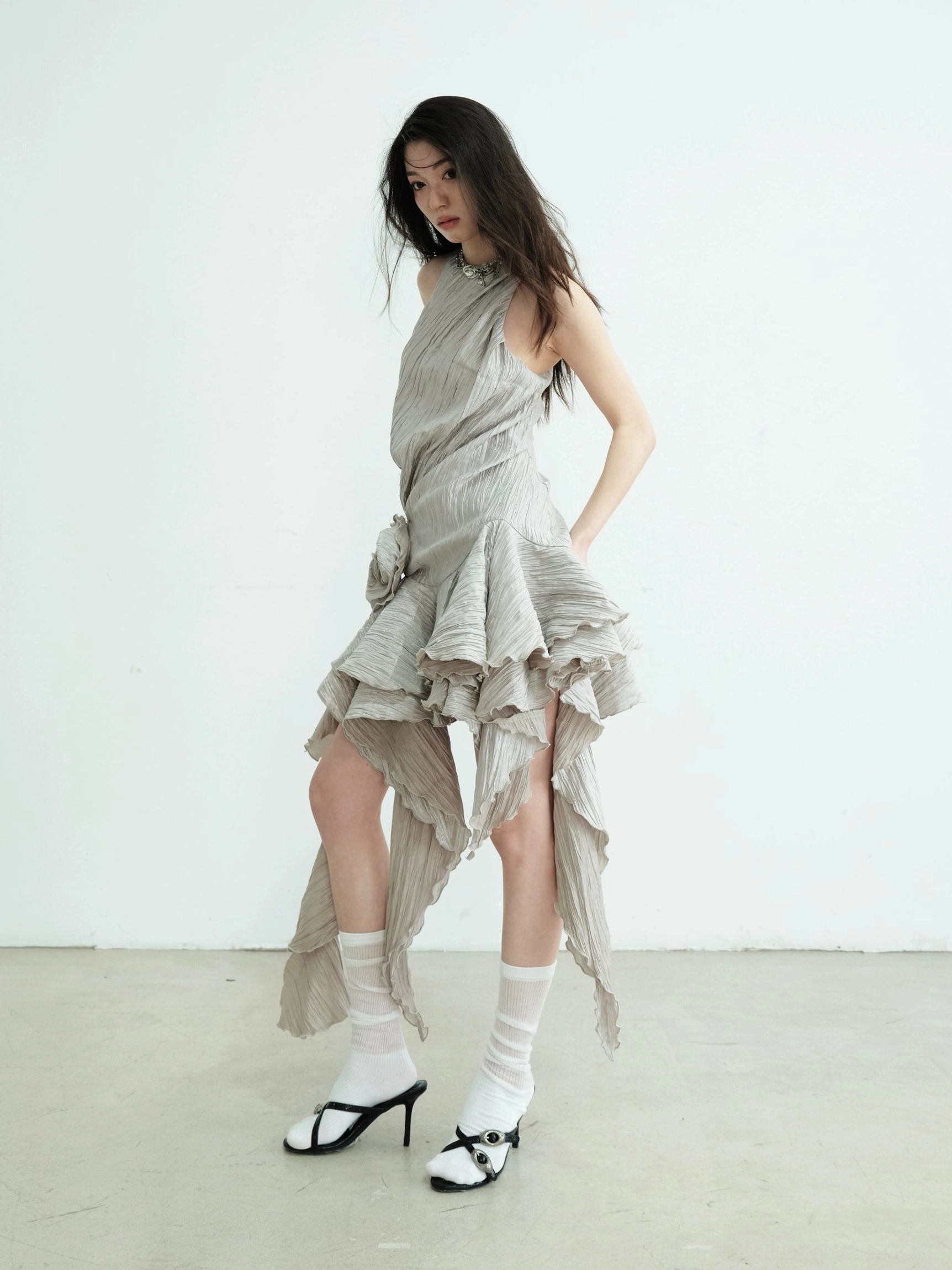 Side view of an asymmetric ruffle hem metallic mini dress featuring a halter neck, draped hem, textured ash fabric, white socks, and bow heels