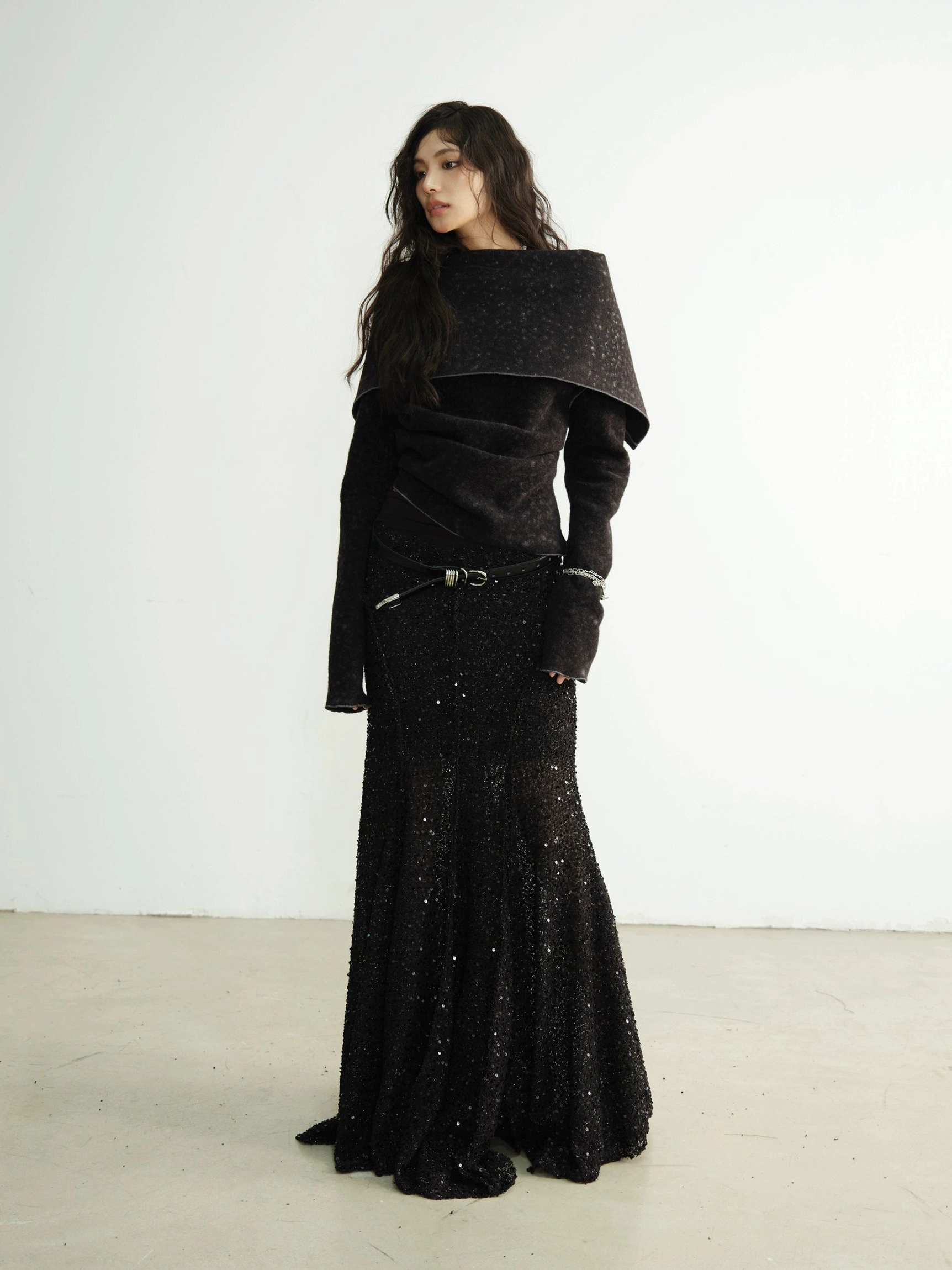 Back three-quarter view of an asymmetric wide-collar black cropped knit cardigan with metal belt and sparkly floor-length mermaid skirt