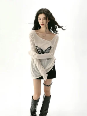 Full body front view of a women's apricot asymmetrical collar butterfly print sun protection long sleeve streetwear top styled with a black mini skirt leg chain and knee high leather boots