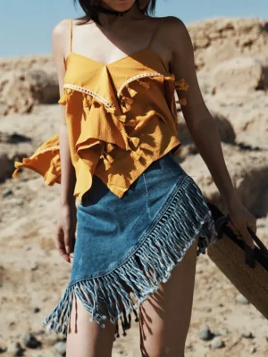 High front three-quarter view of a woman wearing a washed blue asymmetrical fringe denim mini skirt with a yellow ruffled camisole in a desert ruin setting
