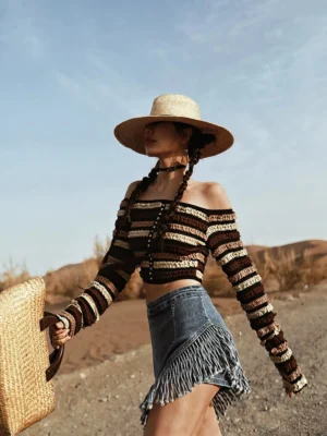 Side view of a woman styling an asymmetrical fringe denim skirt with a striped off-shoulder cropped knit top, straw hat, and woven tote bag