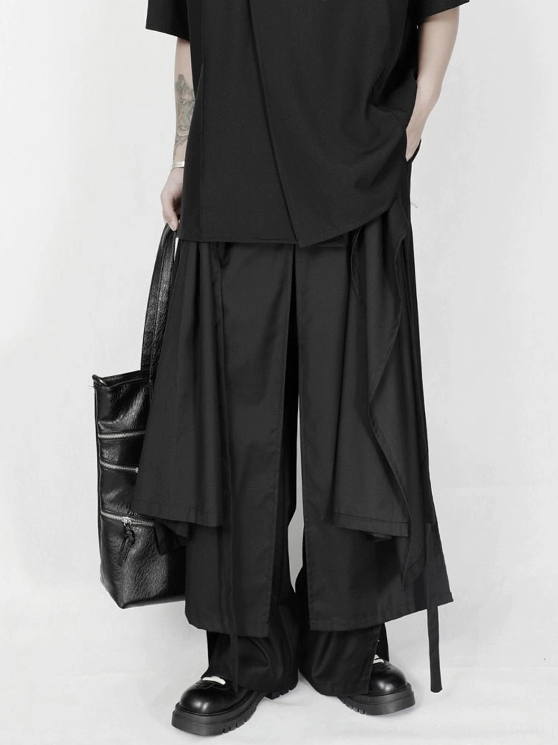 Back three-quarter view of loose black avant-garde wide-leg trousers with asymmetric layers and side hanging straps