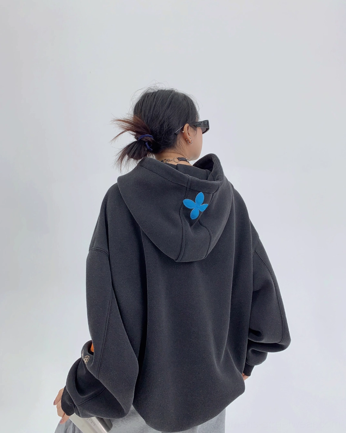 Back view of an oversized black floral hoodie showing the relaxed silhouette and blue flower appliqué detail on the rear of the hood