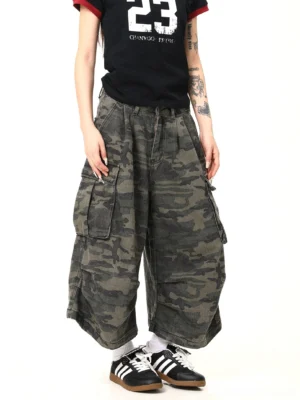 Front angled view of women wearing dark military green camouflage cargo pants with a baggy wide-leg streetwear silhouette, oversized side pockets, pleated high waist, black graphic T-shirt, white socks, and retro training sneakers