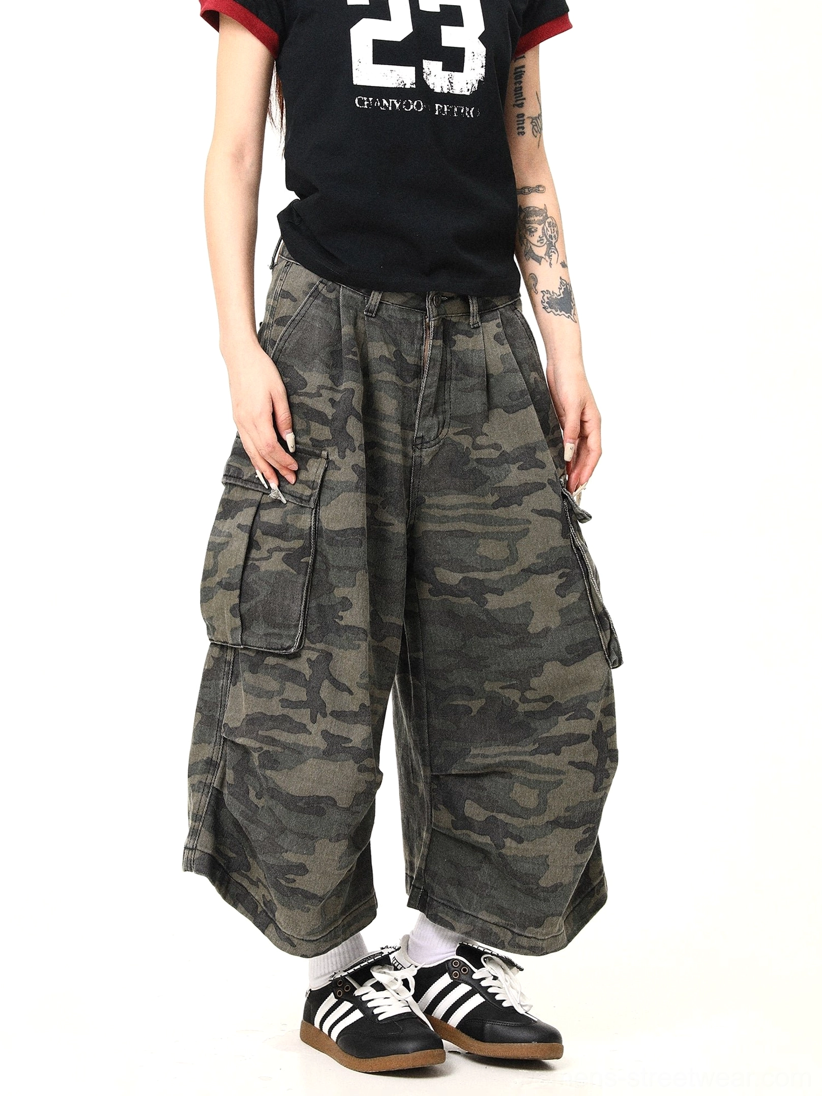 Front angled view of women wearing dark military green camouflage cargo pants with a baggy wide-leg streetwear silhouette, oversized side pockets, pleated high waist, black graphic T-shirt, white socks, and retro training sneakers