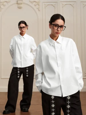 Two models wearing matching white baroque embroidered high-low long-sleeve shirts with wide-leg streetwear trousers in a vintage European studio