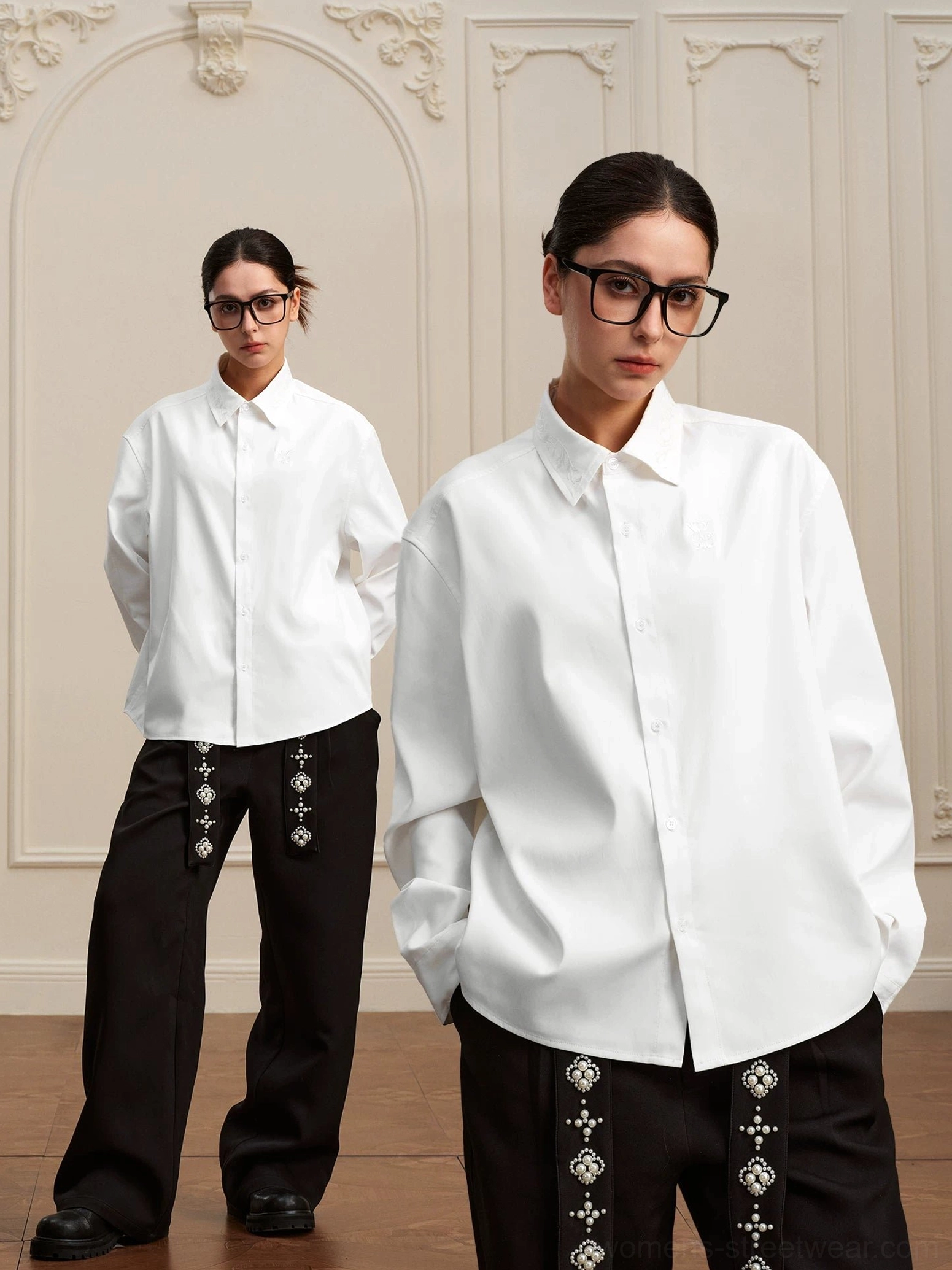 Two models wearing matching white baroque embroidered high-low long-sleeve shirts with wide-leg streetwear trousers in a vintage European studio