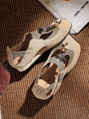 Beige T-strap ballet sandals with striped fabric center straps, decorative beads, and cork footbeds shown from above on a woven rug
