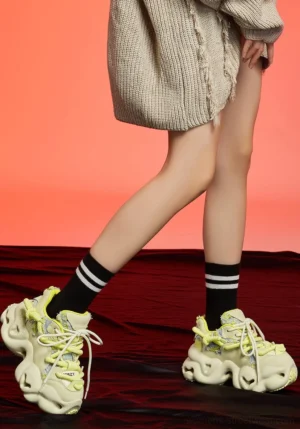 Beige chunky streetwear dad sneakers styled on a model with an oversized knit dress and striped crew socks against a red studio background