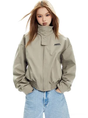 Beige high-collar cropped bomber jacket shown from the front with a relaxed boxy fit, ribbed cuffs, gathered hem, and light blue denim styling