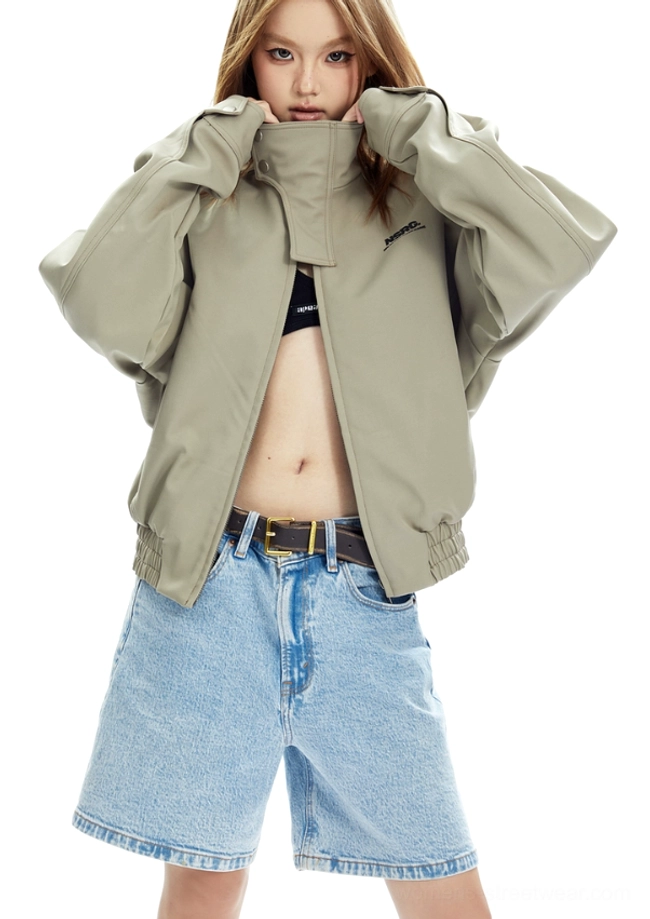Front three-quarter view of a beige high-collar cropped bomber jacket styled over a black crop top with light blue denim