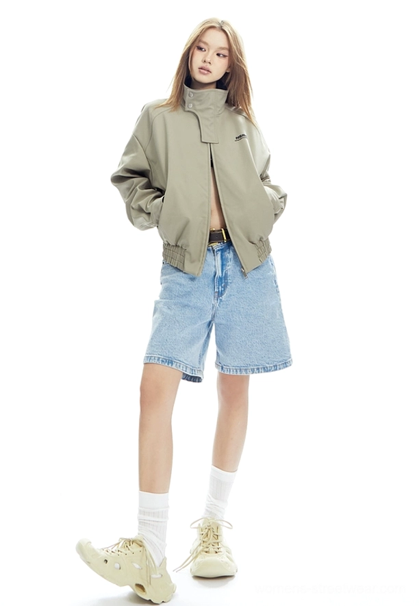 Side-front streetwear styling of a beige high-collar cropped bomber jacket with exposed midriff, denim shorts, and chunky sneakers