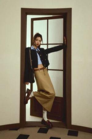 Side view of a woman wearing a beige khaki maxi skirt with a brown leather belt and loafers beside a wooden glass door