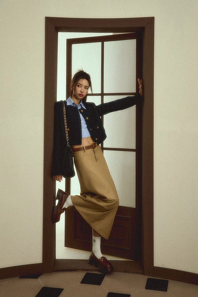 Side view of a woman wearing a beige khaki maxi skirt with a brown leather belt and loafers beside a wooden glass door