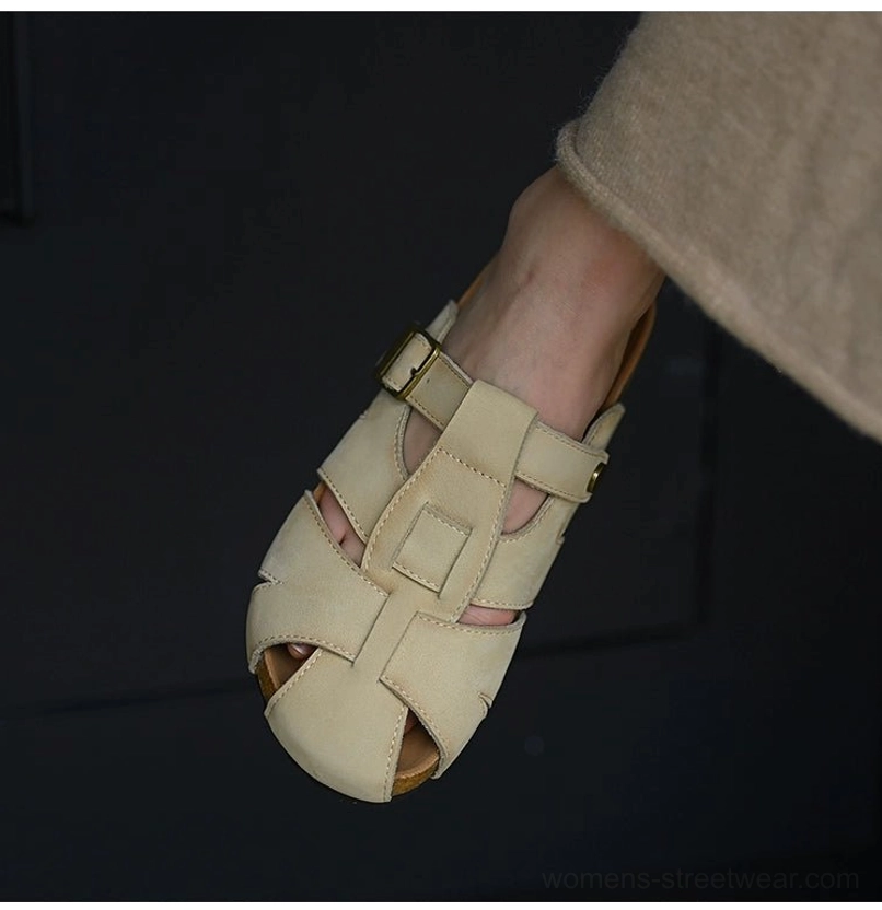 Low-angle view of beige leather fisherman sandals styled with trousers, featuring a rounded closed toe, multi-cutout straps, adjustable side buckle, and cork footbed