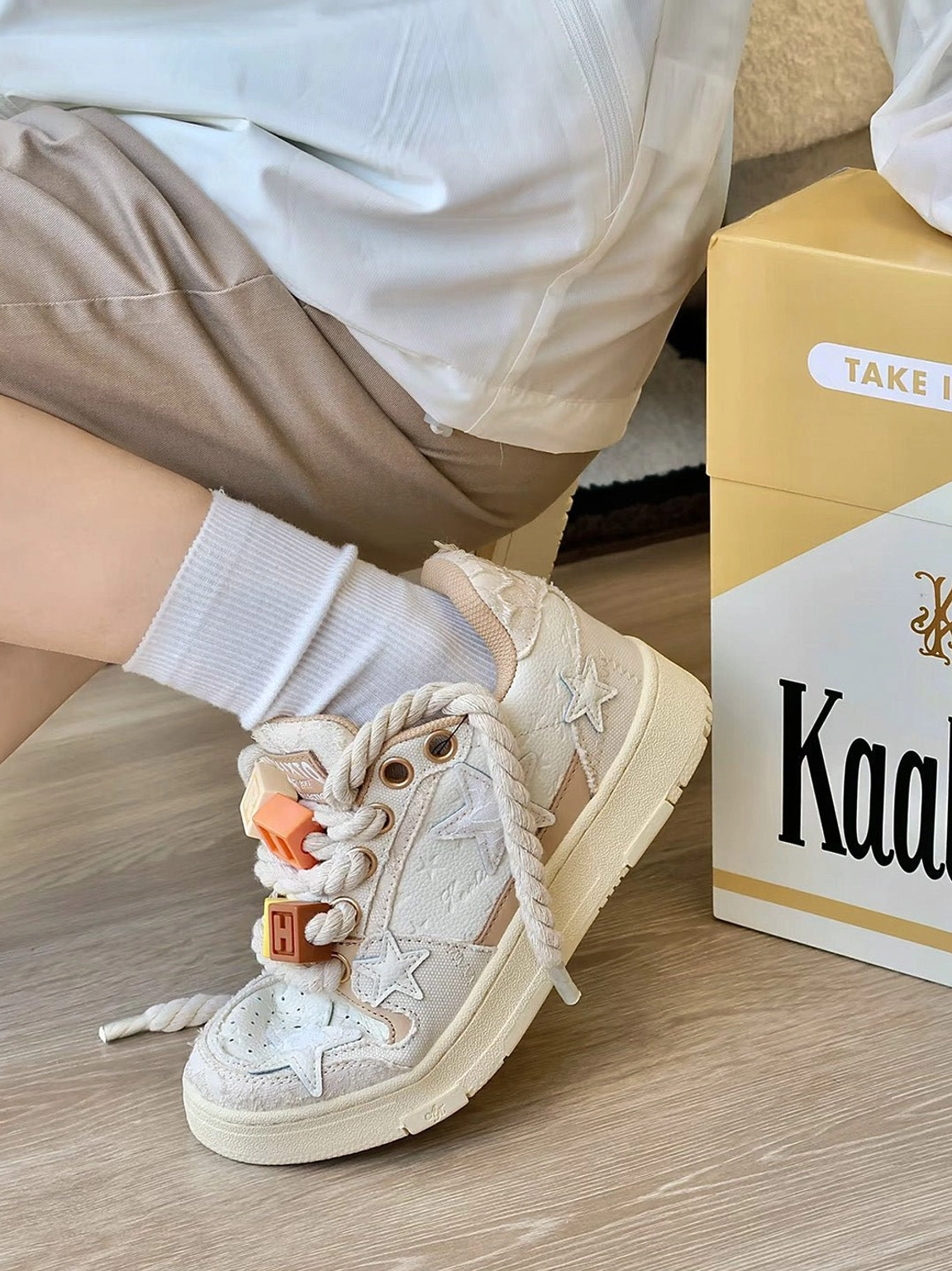 Beige low-top star sneakers worn with white socks and khaki shorts, featuring colorful rope lace charms and retro streetwear detailing