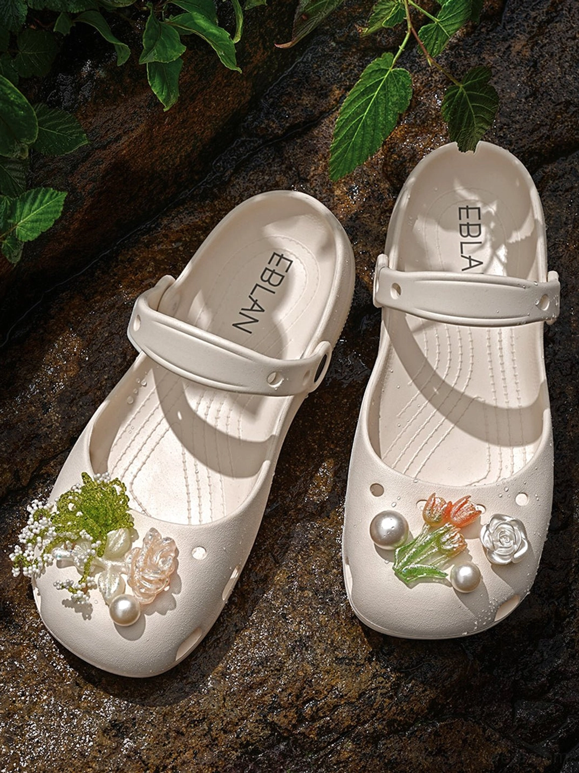 Beige Mary Jane slide sandals displayed on dark rock with green foliage showing hollow upper floral pearl details and lifted sole