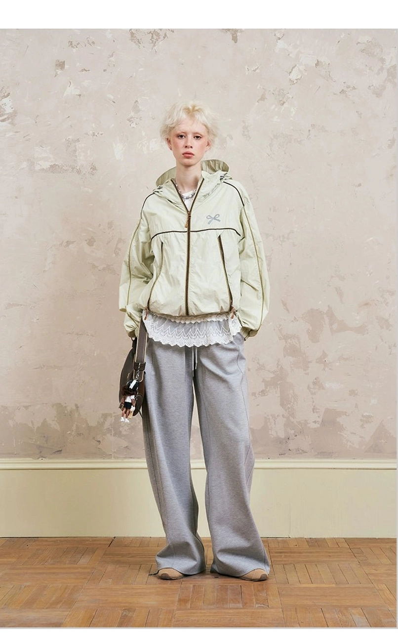 Front view of beige sporty hooded jacket with brown contrast piping, half-zip closure, bow embroidery, gray wide-leg sweatpants, and handheld leather bag
