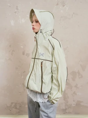 Side view of beige sporty hooded windbreaker jacket with contrast piping, half-zip front, adjustable hood and hem, gray wide-leg pants, and lace underlayer