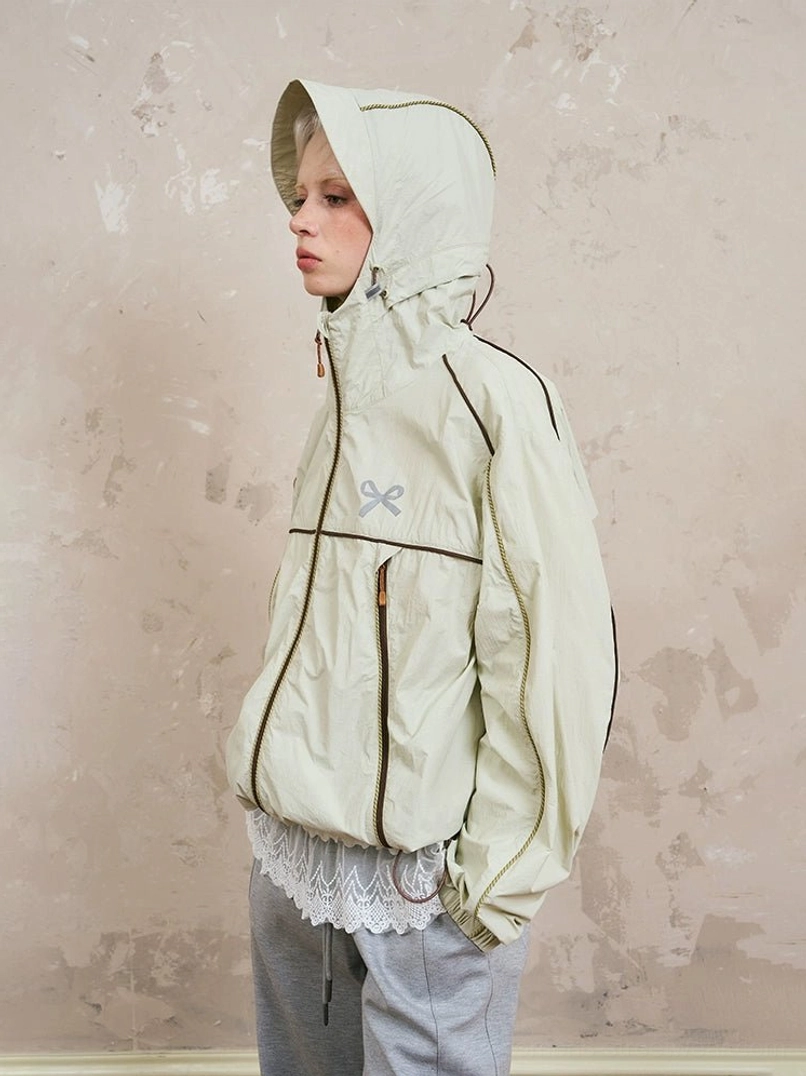 Side view of beige sporty hooded windbreaker jacket with contrast piping, half-zip front, adjustable hood and hem, gray wide-leg pants, and lace underlayer