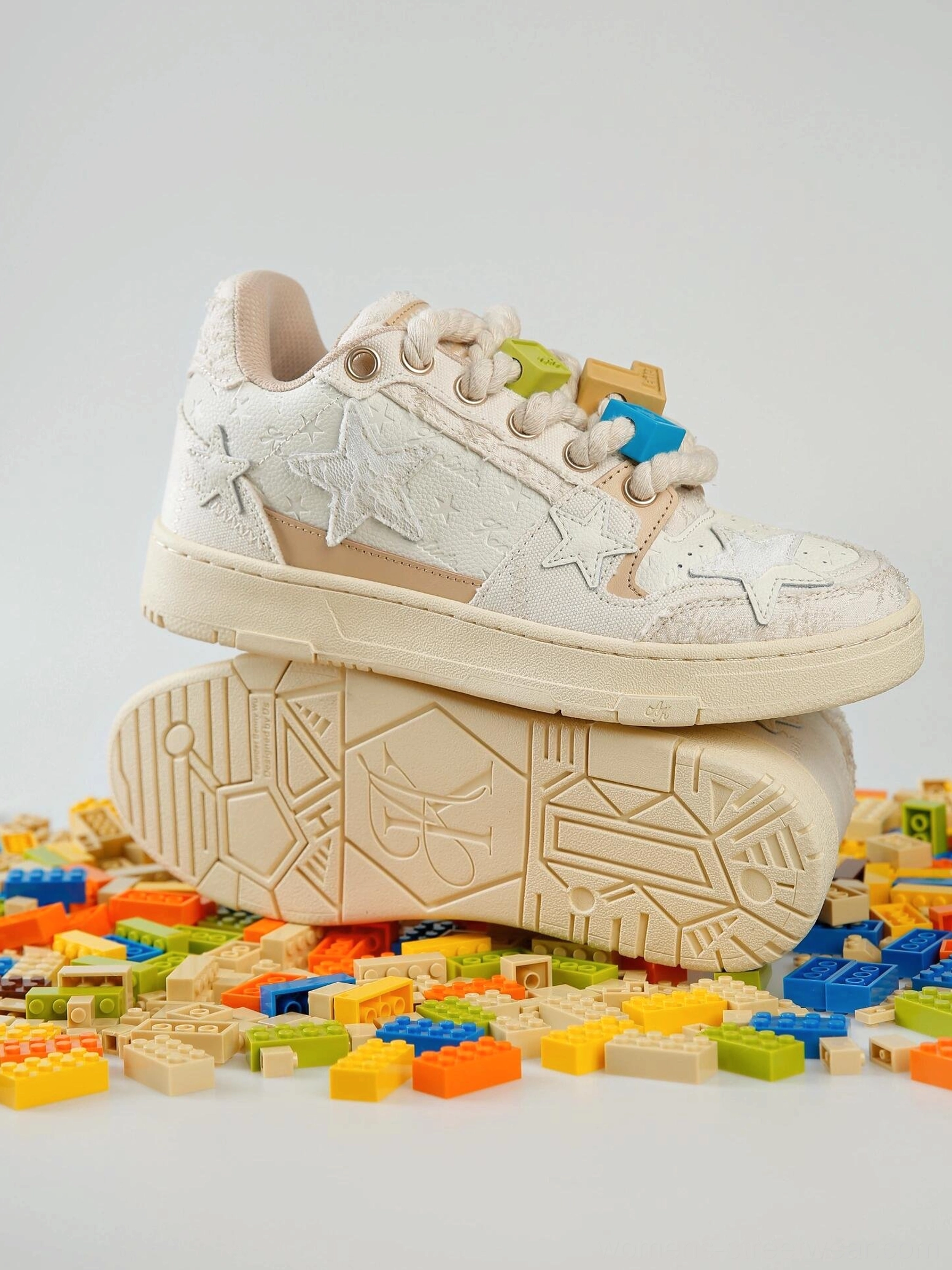 Beige star low-top sneakers stacked to show the textured upper, decorative lace charms, thick rubber outsole, and distressed streetwear finish