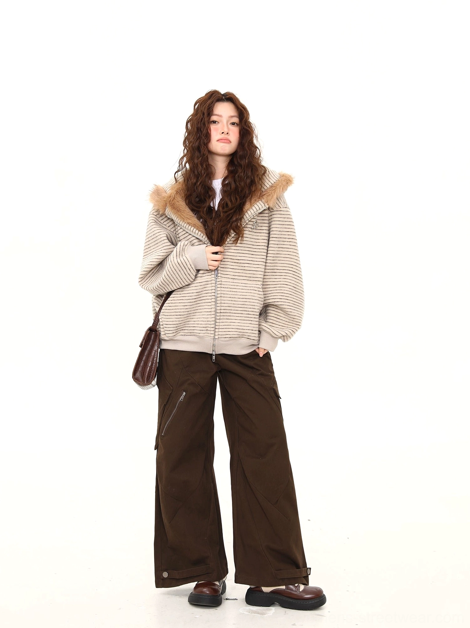Three-quarter styling view of a beige striped bunny-ear zip-up hoodie with subtle chest embroidery, shoulder bag, and wide-leg pants
