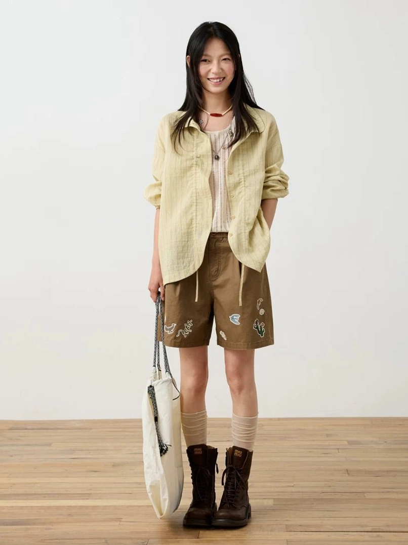 Beige striped textured button-up shirt and camisole styled with embroidered brown shorts, socks, combat boots, and a canvas tote