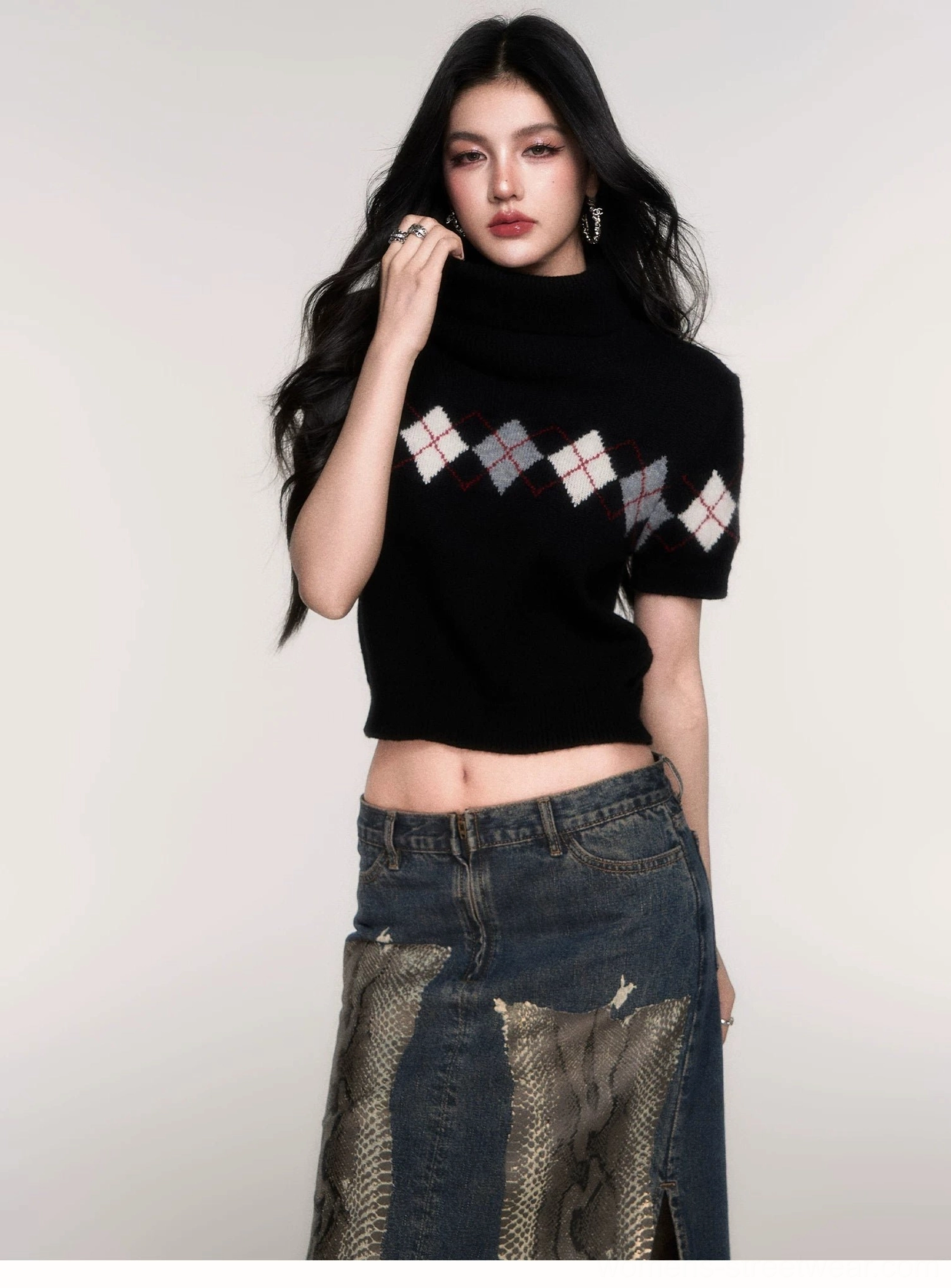 Front three-quarter view of a black argyle cropped turtleneck sweater with short sleeves and gray white wine-red diamond knit panel styled with a patchwork denim midi skirt