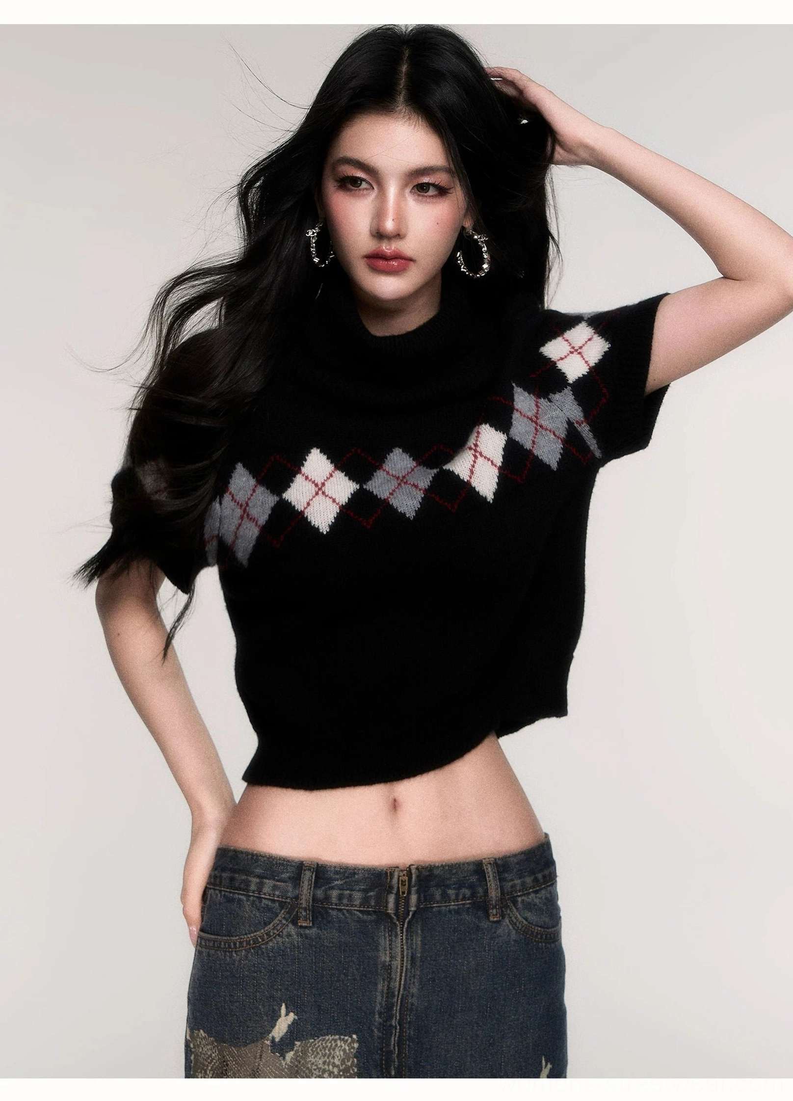 Side front view of a black argyle cropped turtleneck sweater showing the exposed waist styling with low-rise denim jeans and hoop earrings