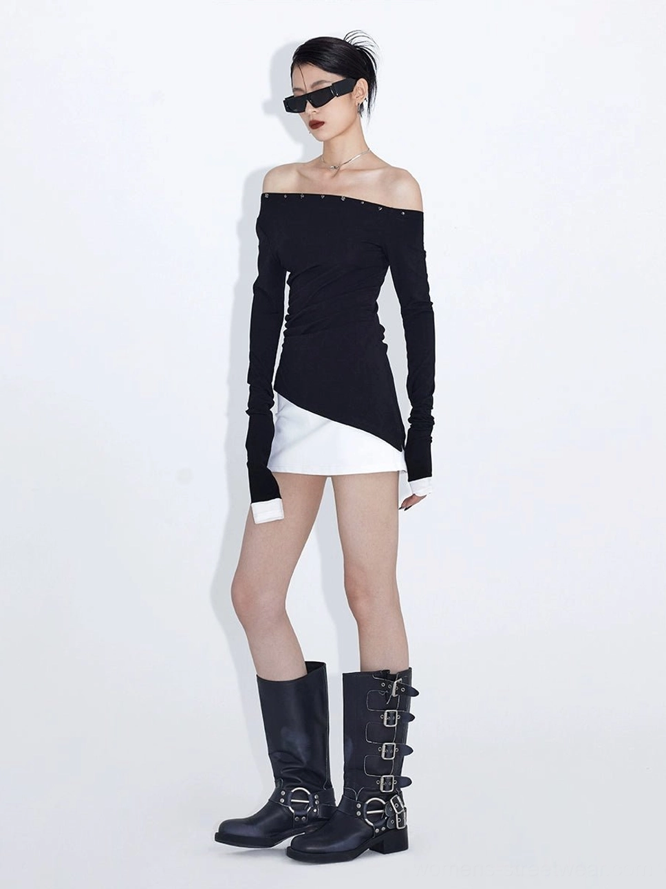 Front full-body styling of a black asymmetric off-shoulder top with extra-long sleeves, studded neckline, white mini skirt, sunglasses, and mid-calf buckle boots