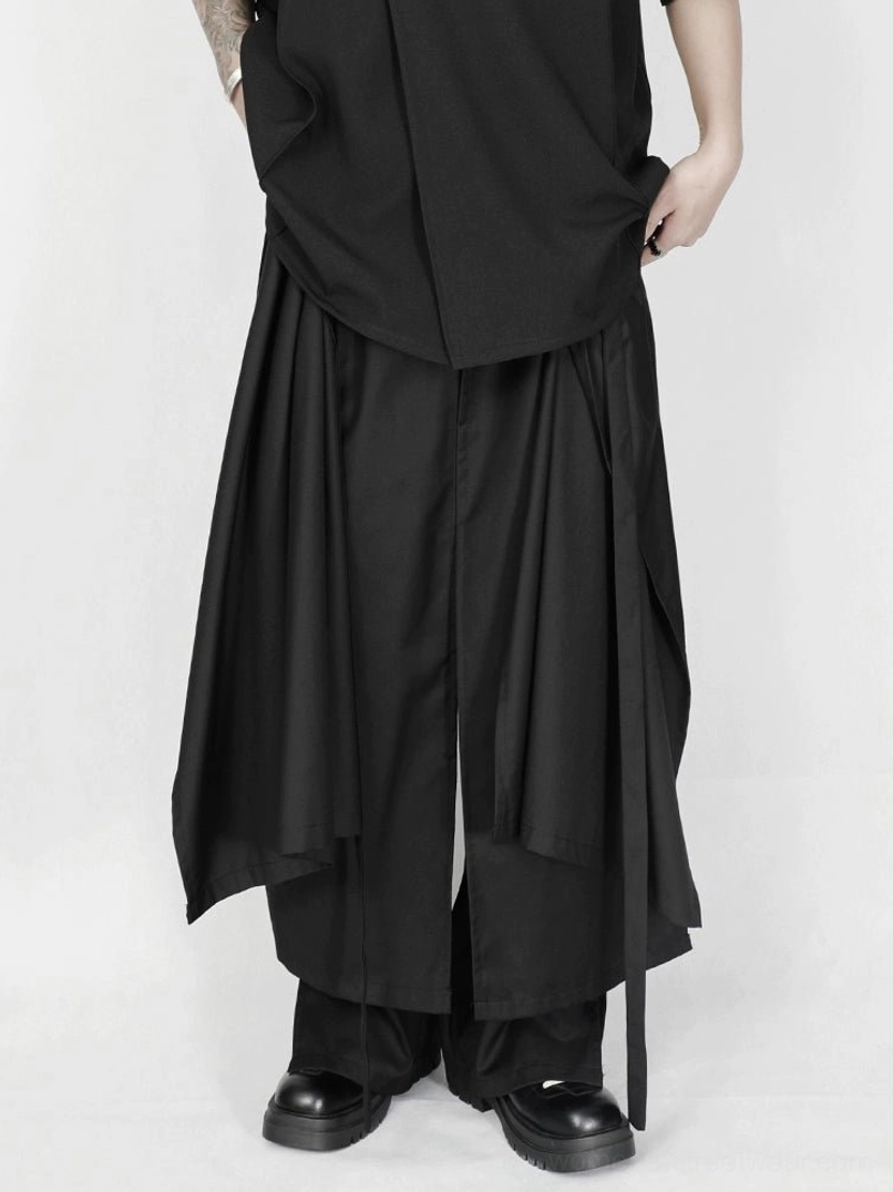 Back lower-body detail of black asymmetric layered wide-leg trousers with a hanging strap at the hem