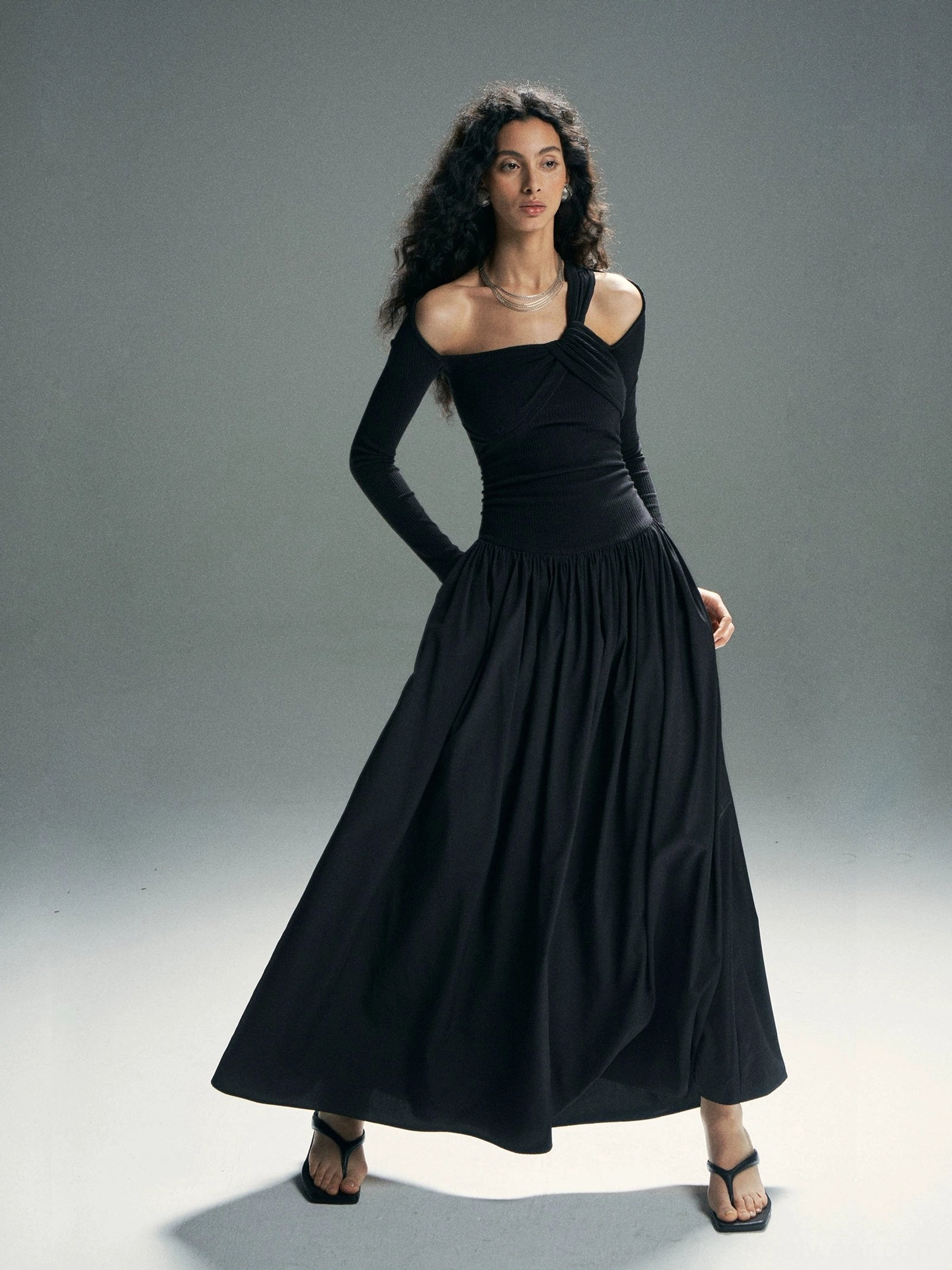 Black asymmetrical cold-shoulder maxi dress shown from the front three-quarter angle with a fitted bodice, long sleeve, and gathered floor-length skirt