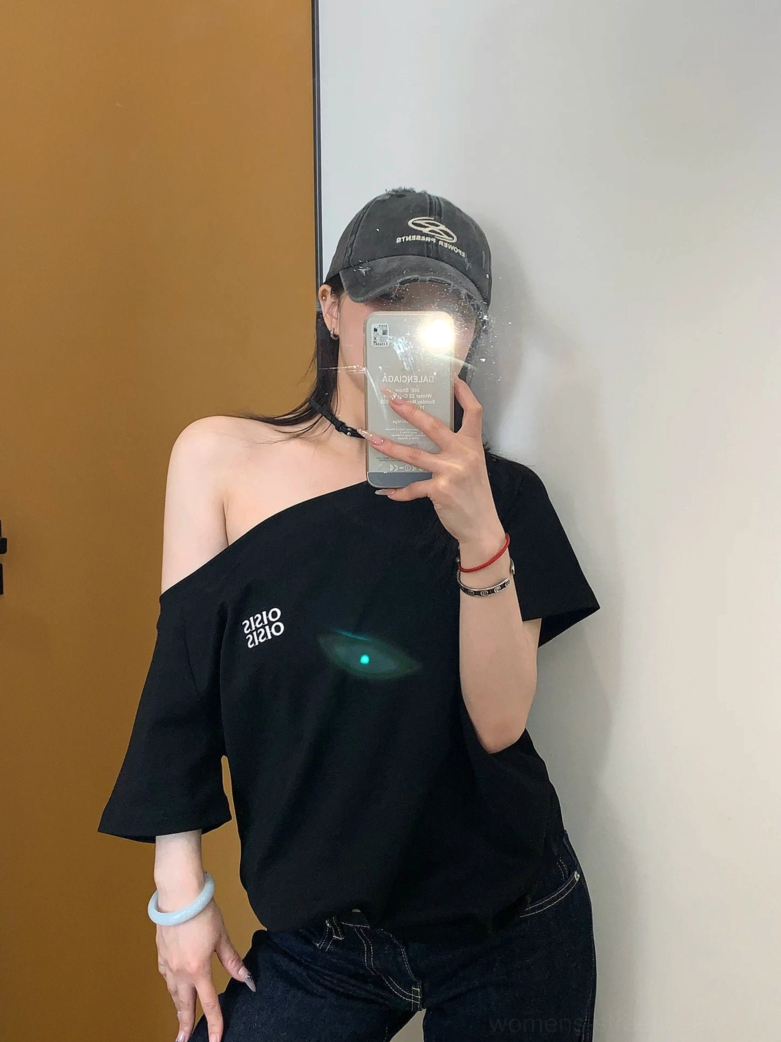 Front view of a black asymmetrical off-shoulder logo tee with white chest print styled with dark denim and a baseball cap