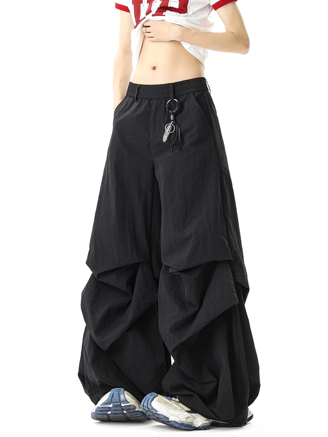 Front three-quarter view of black avant-garde parachute streetwear pants with layered ruched legs, elastic waistband, detachable key ring detail, cropped top, and chunky sneakers