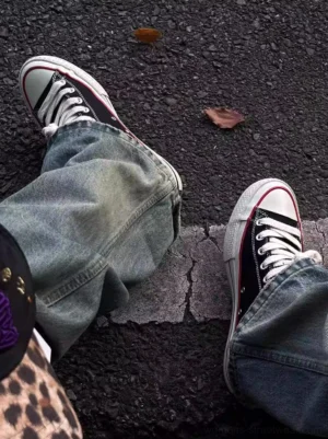 Black bat print canvas sneakers worn with loose blue denim jeans on an urban asphalt street with fallen leaves, showing red trim streetwear styling